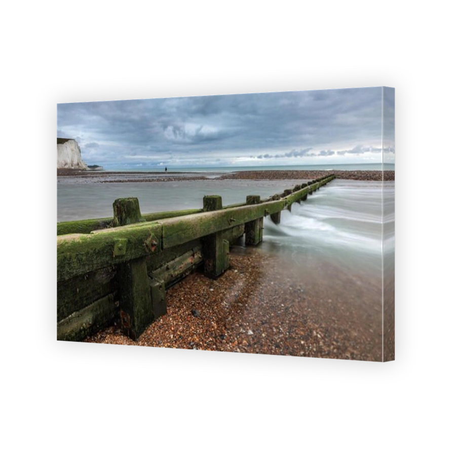 Wooden beach groyne, covered in green seaweed extending into the sea, lone figure in distance, at Cuckmere Haven, Seaford, East Sussex, England, UK