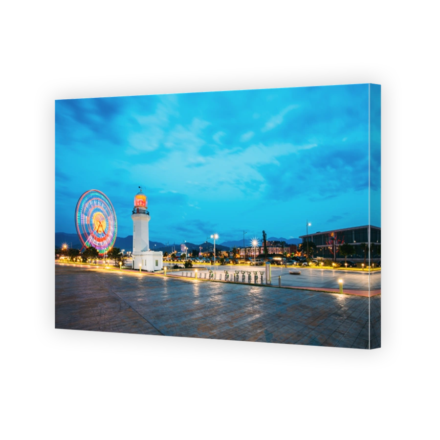 Batumi, Adjara, Georgia. Ferris Wheel In Motion And Pitsunda Lighthouse At Promenade In Miracle Park, Amusement City Park On Blue Evening Sky And Hilly Background. Blue Hour. Night Time