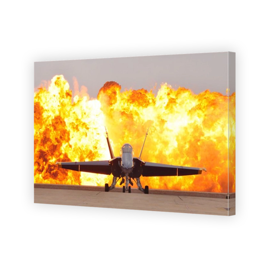 An F/A-18 Hornet sits on the flight line as a wall of fire detonates behind it.